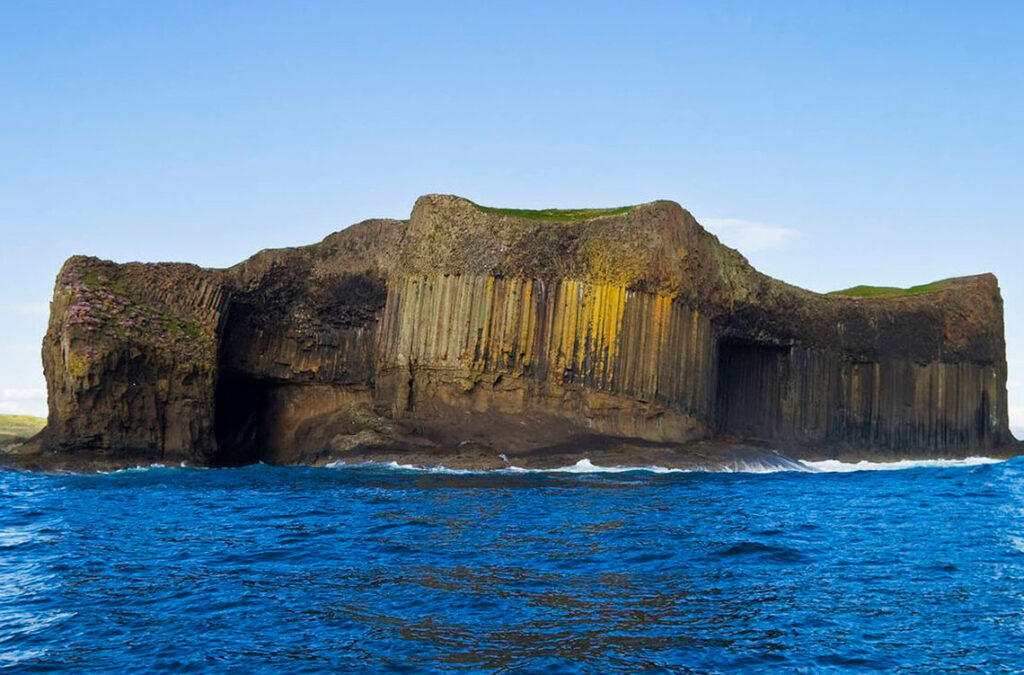 Vista distante de la Cueva de Fingal en Escocia, emergiendo imponente desde el mar. Esta maravilla geológica, visible incluso desde lejos, invita a los visitantes a descubrir sus secretos ancestrales y formaciones únicas. / Foto: iStock